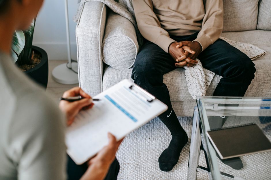 Blurred unrecognizable female therapist taking notes on clipboard while sitting in front of ethnic male patient with clasped hands during meeting